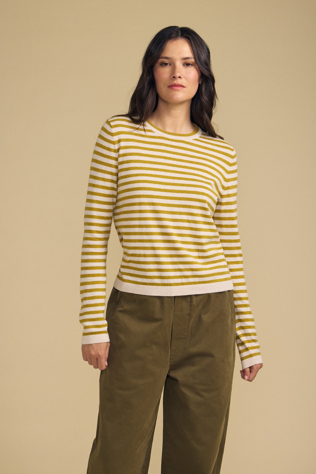 A person stands against a beige background wearing the ROXY SWEATER by Velvet by Graham & Spencer—a yellow and white striped sweater—paired with olive-green pants.