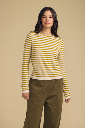 A person stands against a beige background wearing the ROXY SWEATER by Velvet by Graham & Spencer—a yellow and white striped sweater—paired with olive-green pants.