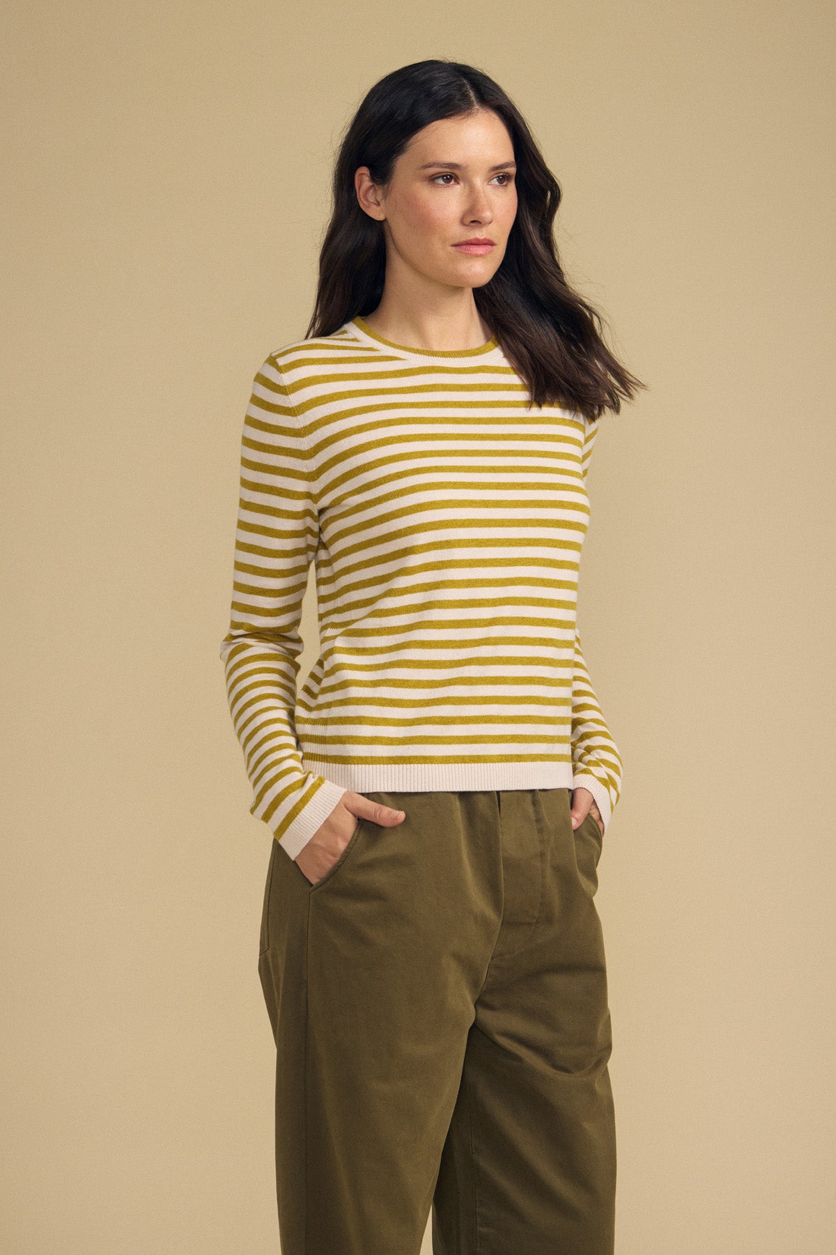 A woman stands against a beige background wearing the ROXY SWEATER by Velvet by Graham & Spencer—a yellow and white striped sweater—paired with olive green pants, her hands in her pockets.