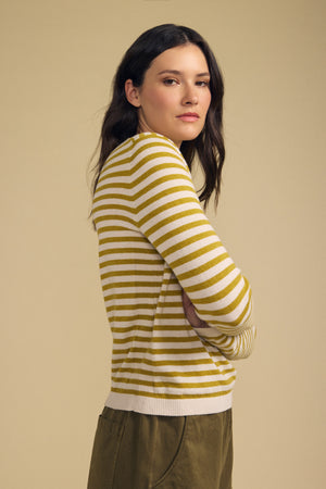 A woman with long dark hair stands sideways against a beige background, arms crossed and looking at the camera, wearing the ROXY SWEATER by Velvet by Graham & Spencer in yellow and white stripes paired with olive pants.