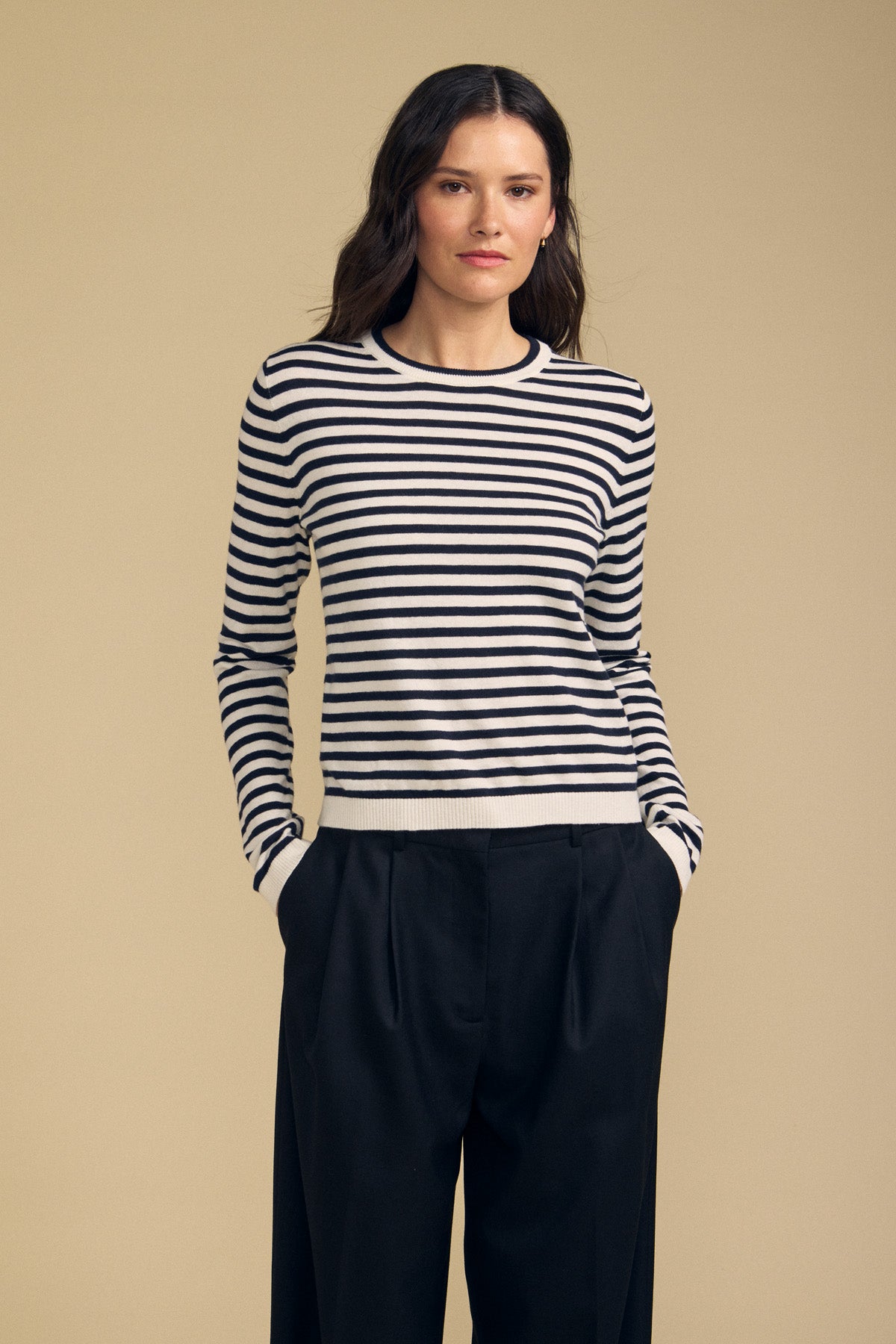 Standing against a simple beige backdrop, the individual wears the ROXY SWEATER by Velvet by Graham & Spencer—a black and white striped piece crafted from a soft cotton-cashmere blend—paired with black pants and hands tucked into pockets.