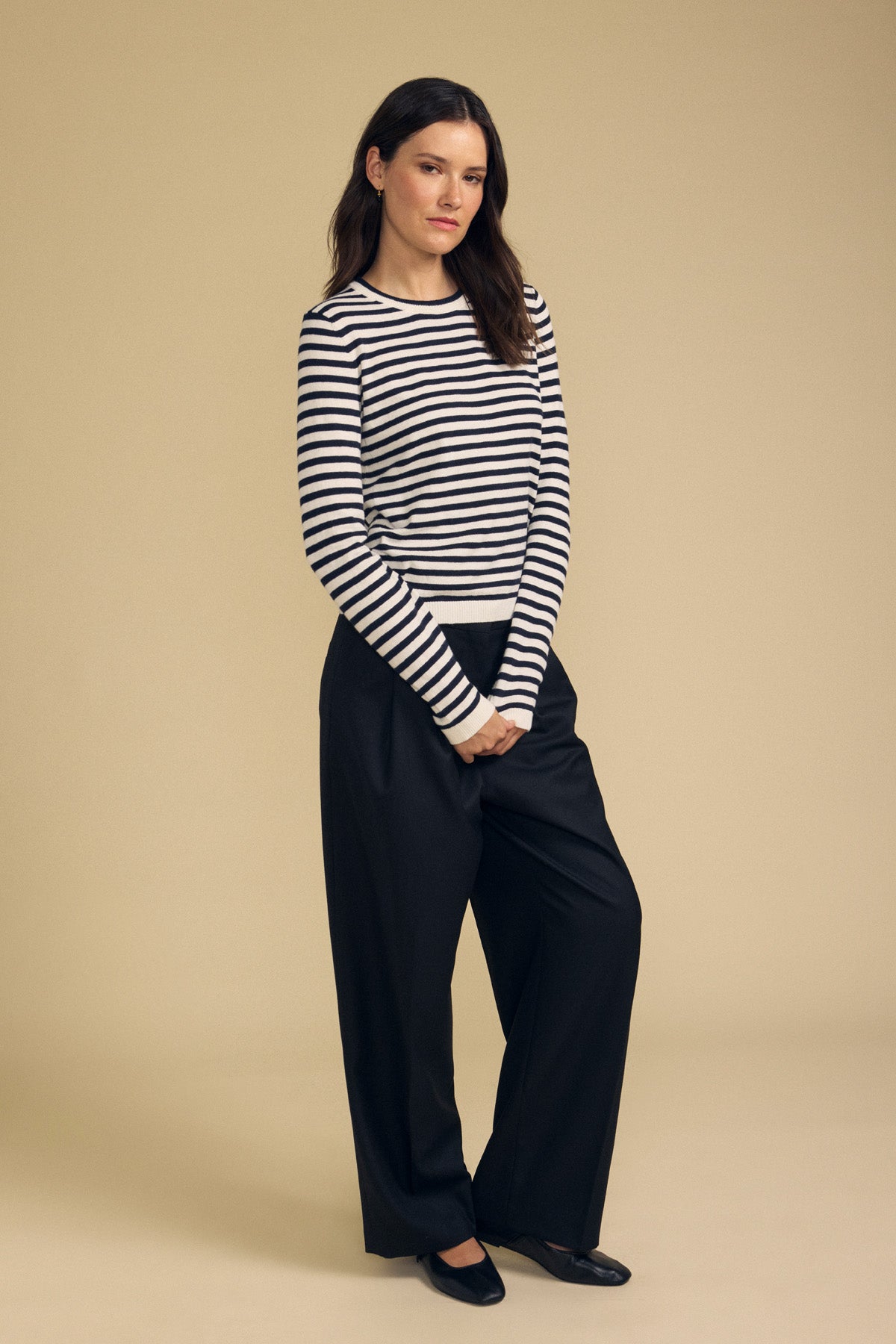 A woman stands against a beige background wearing the ROXY SWEATER by Velvet by Graham & Spencer—a black and white striped sweater crafted from a lightweight cotton-cashmere blend—paired with black wide-leg pants and black shoes, her hands clasped in front.-41965253492929