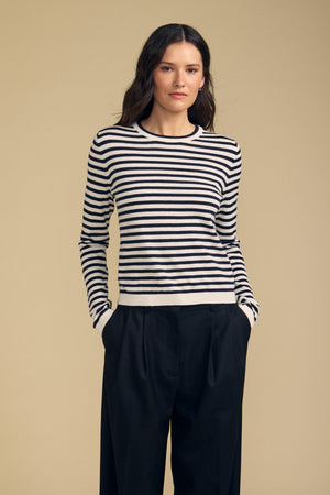 Standing against a simple beige backdrop, the individual wears the ROXY SWEATER by Velvet by Graham & Spencer—a black and white striped piece crafted from a soft cotton-cashmere blend—paired with black pants and hands tucked into pockets.