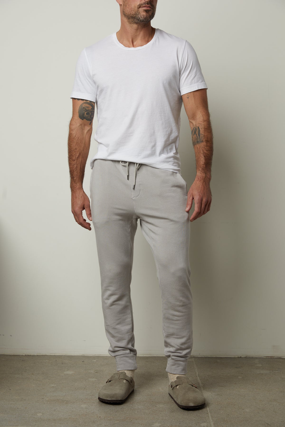   A person wearing a white t-shirt, Crosby Jogger from Velvet by Graham & Spencer in gray luxe fleece knit, and beige slippers stands against a plain wall. 