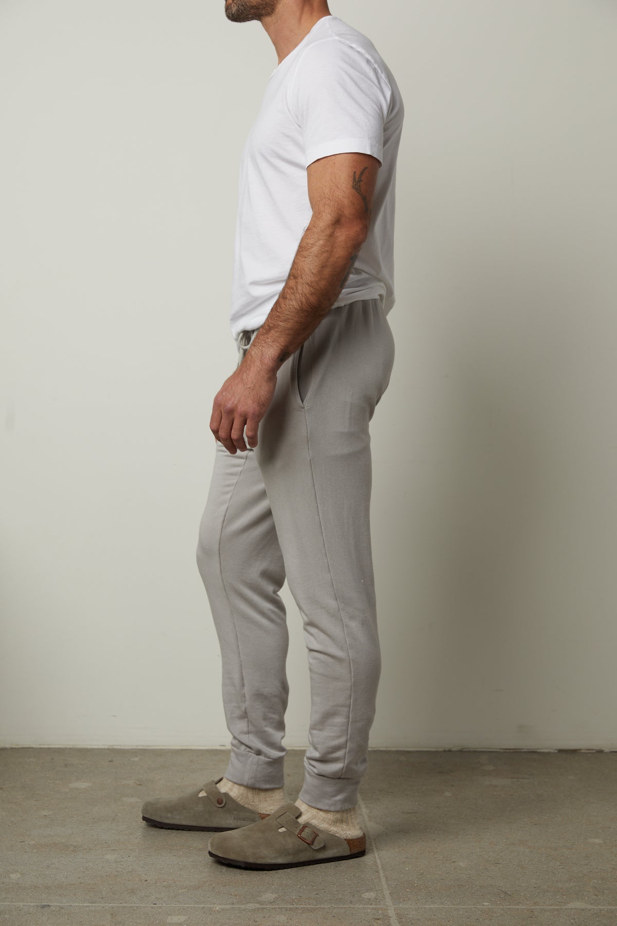   A person wearing a white t-shirt, the CROSBY JOGGER from Velvet by Graham & Spencer made of luxe fleece knit in versatile gray, and beige slippers stands against a plain light gray background. 