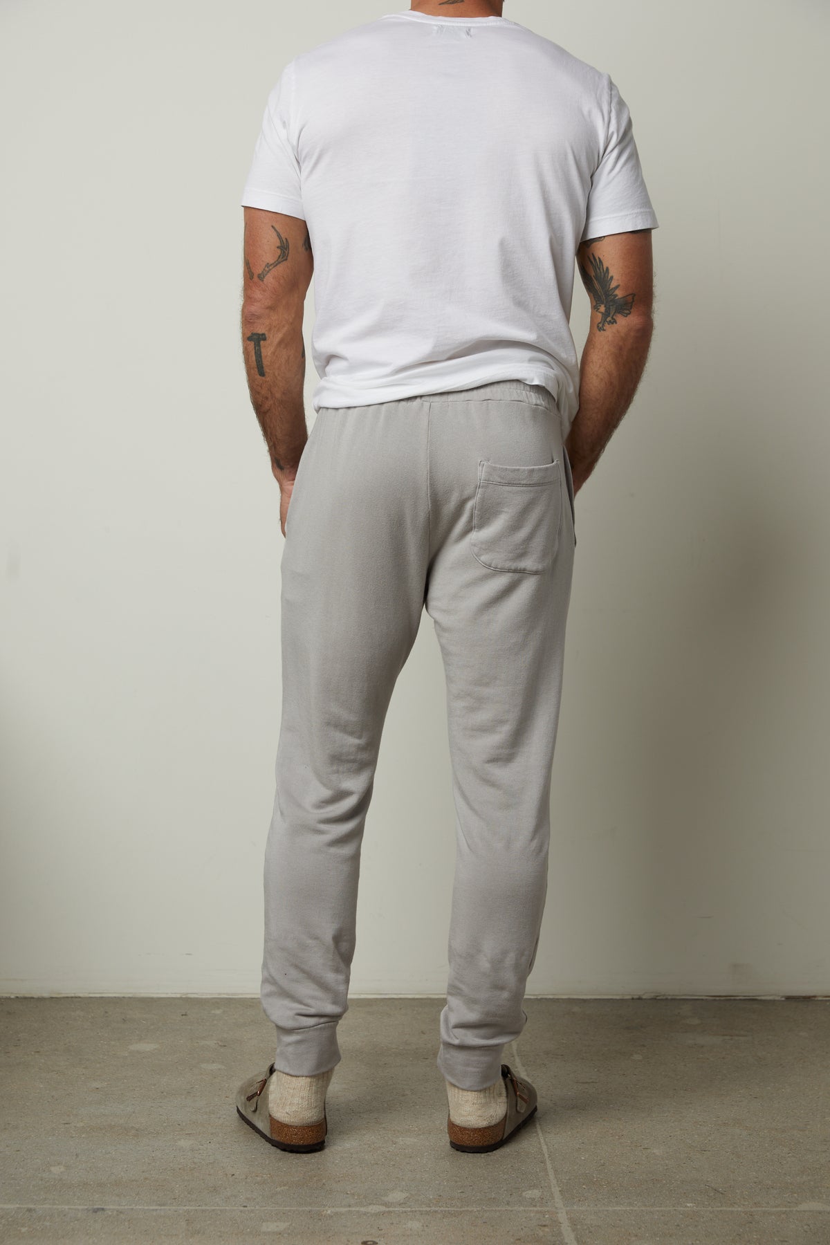   A person in a white t-shirt and the CROSBY JOGGER by Velvet by Graham & Spencer stands facing away, showcasing their tattooed arms while wearing sandals. The simple wall and floor backdrop perfectly highlight the versatile style of their luxe fleece knit ensemble. 