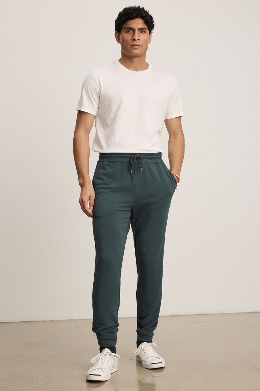 A man stands against a plain background wearing a white t-shirt, white sneakers, and dark green Velvet by Graham & Spencer CROSBY JOGGER pants, with his hands in his pockets.