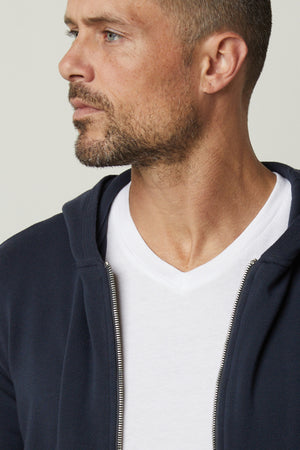 A man wearing a Velvet by Graham & Spencer navy RODAN LUXE FLEECE ZIP HOODIE and white t-shirt, showcasing the intricate fabrication details reminiscent of gym-class attire.