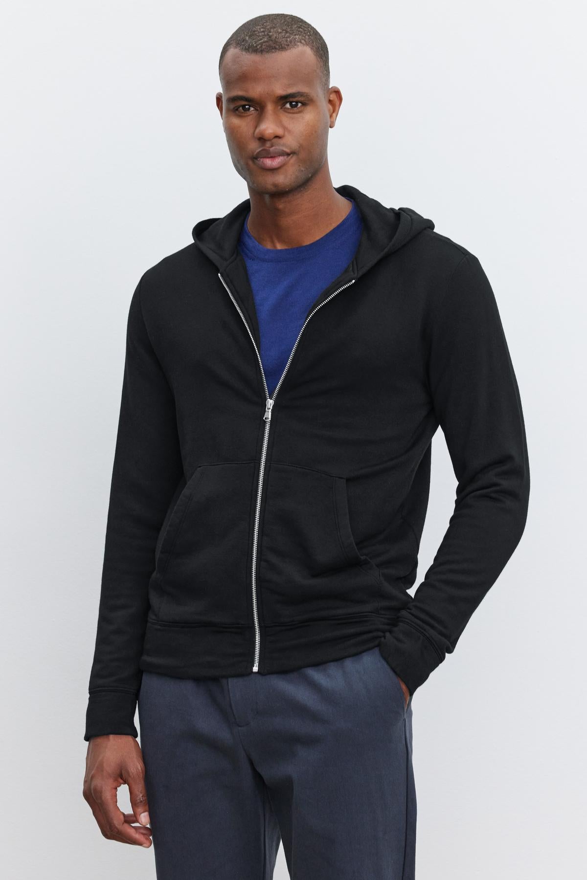   A person stands against a plain background wearing the RODAN HOODIE by Velvet by Graham & Spencer over a blue shirt and grey pants. 