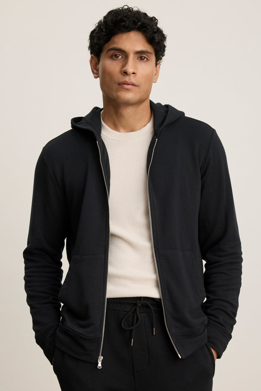 A man with short curly hair stands with his hands in the pockets of a RODAN HOODIE by Velvet by Graham & Spencer, layered over a light beige shirt and black pants, against a plain light background.