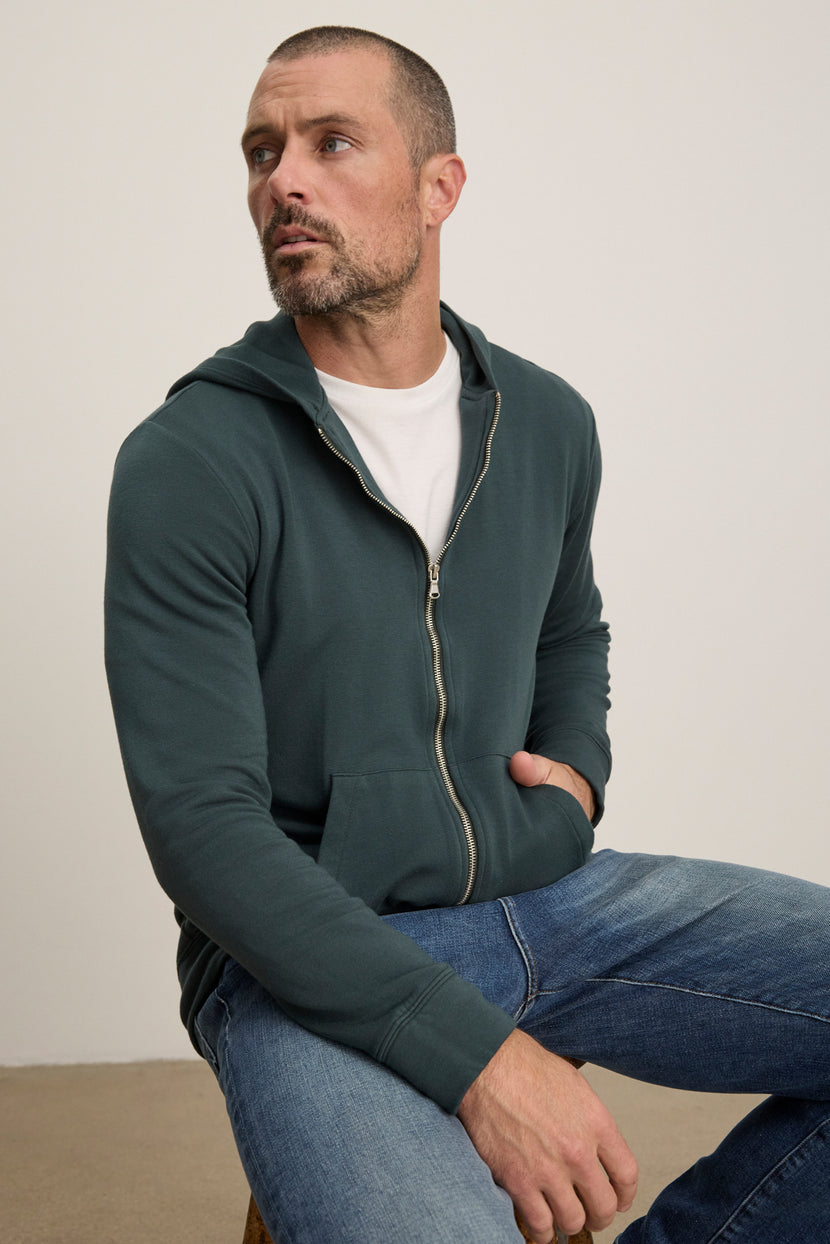 A man with short hair and a beard sits on a stool against a plain background, wearing the RODAN HOODIE by Velvet by Graham & Spencer over a white t-shirt and blue jeans.