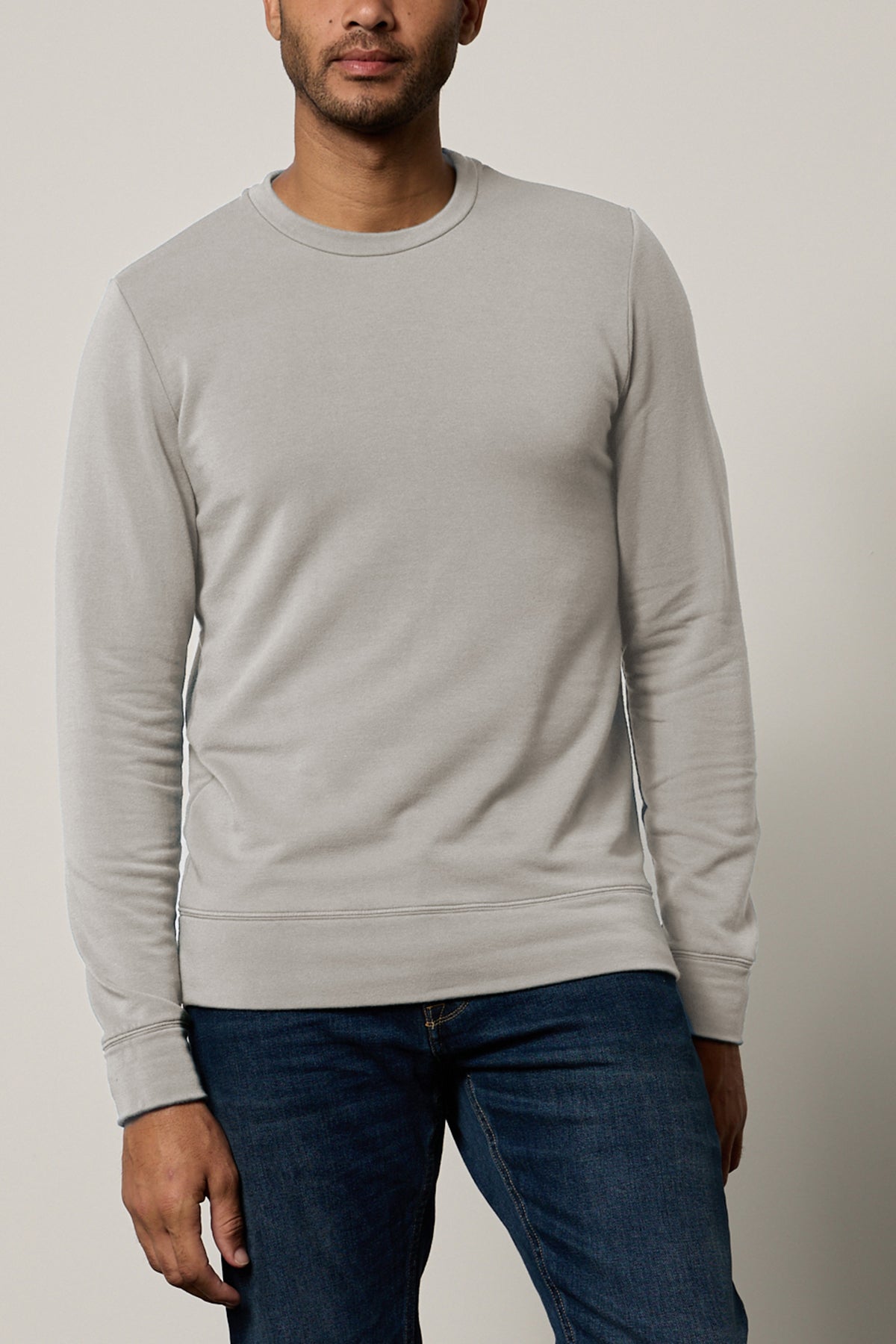   A person wearing a light grey SOREN PULLOVER by Velvet by Graham & Spencer and blue jeans stands against a plain backdrop. 