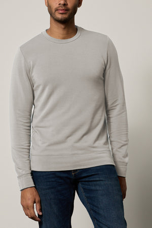 A person wearing a light grey SOREN PULLOVER by Velvet by Graham & Spencer and blue jeans stands against a plain backdrop.