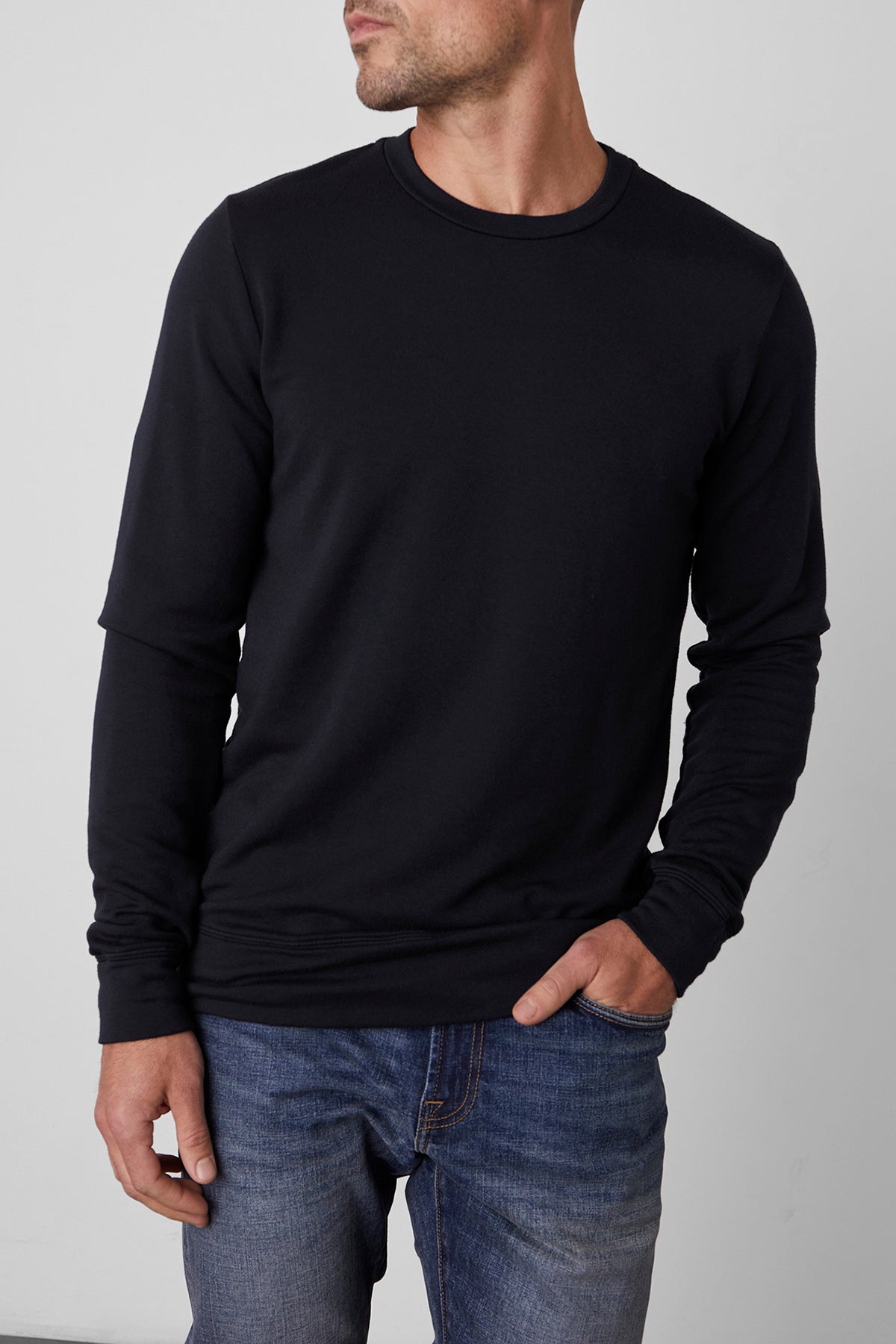   A person wearing the SOREN PULLOVER from Velvet by Graham & Spencer, a black long-sleeved shirt with a cozy brushed interior, along with blue jeans stands with one hand in their pocket, facing slightly to the side. 