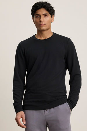 A person with short, curly dark hair wears the SOREN PULLOVER by Velvet by Graham & Spencer, featuring a cozy brushed interior, paired with light gray pants and standing against a neutral background.
