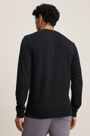 A person is seen from behind wearing the SOREN PULLOVER by Velvet by Graham & Spencer, a plain black luxe fleece with a cozy brushed interior, paired with gray pants and standing against a neutral background.