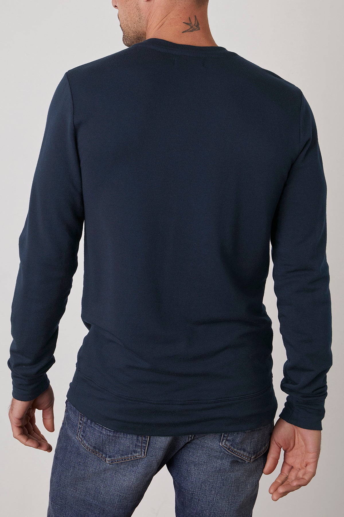   An individual wearing the SOREN PULLOVER by Velvet by Graham & Spencer, paired with jeans, is shown from behind, revealing a small tattoo on their neck. 