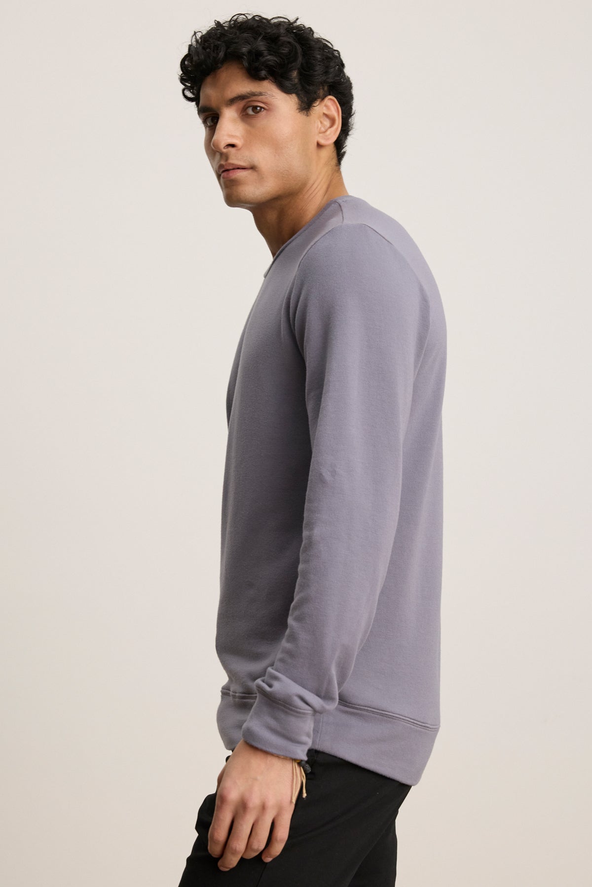 A man with short curly hair, in black pants and a light purple SOREN PULLOVER by Velvet by Graham & Spencer, stands sideways against a plain background.-39841104789697