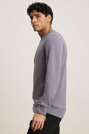 A man with short curly hair, in black pants and a light purple SOREN PULLOVER by Velvet by Graham & Spencer, stands sideways against a plain background.
