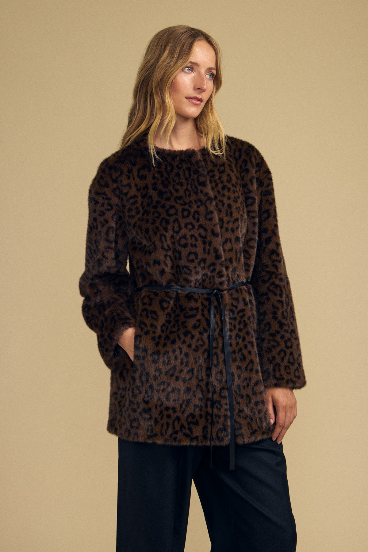 A woman wearing the MARMONT FAUX FUR JACKET by Velvet by Graham & Spencer—a brown leopard print jacket featuring snap button closures and a black belt—stands against a plain beige background.-42063811739841