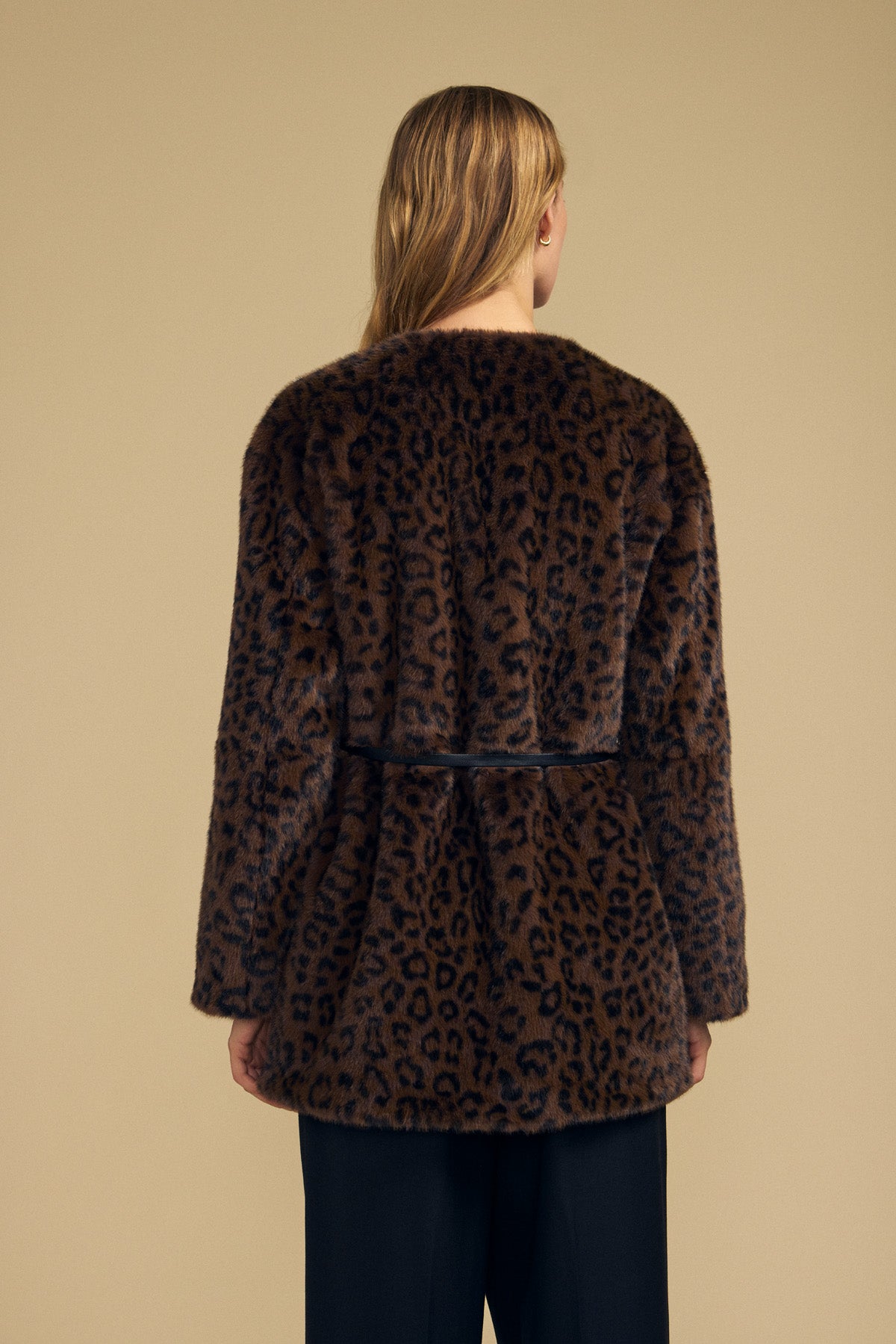 A person with long blonde hair is seen from behind wearing the Velvet by Graham & Spencer MARMONT FAUX FUR JACKET, a brown leopard-print style featuring snap button closures, paired with black pants against a beige background.-42063811379393