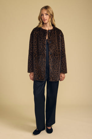 A person stands facing forward against a plain beige background, wearing the MARMONT FAUX FUR JACKET by Velvet by Graham & Spencer—a brown leopard-print faux fur jacket with snap button closures—styled over a black top and black pants.