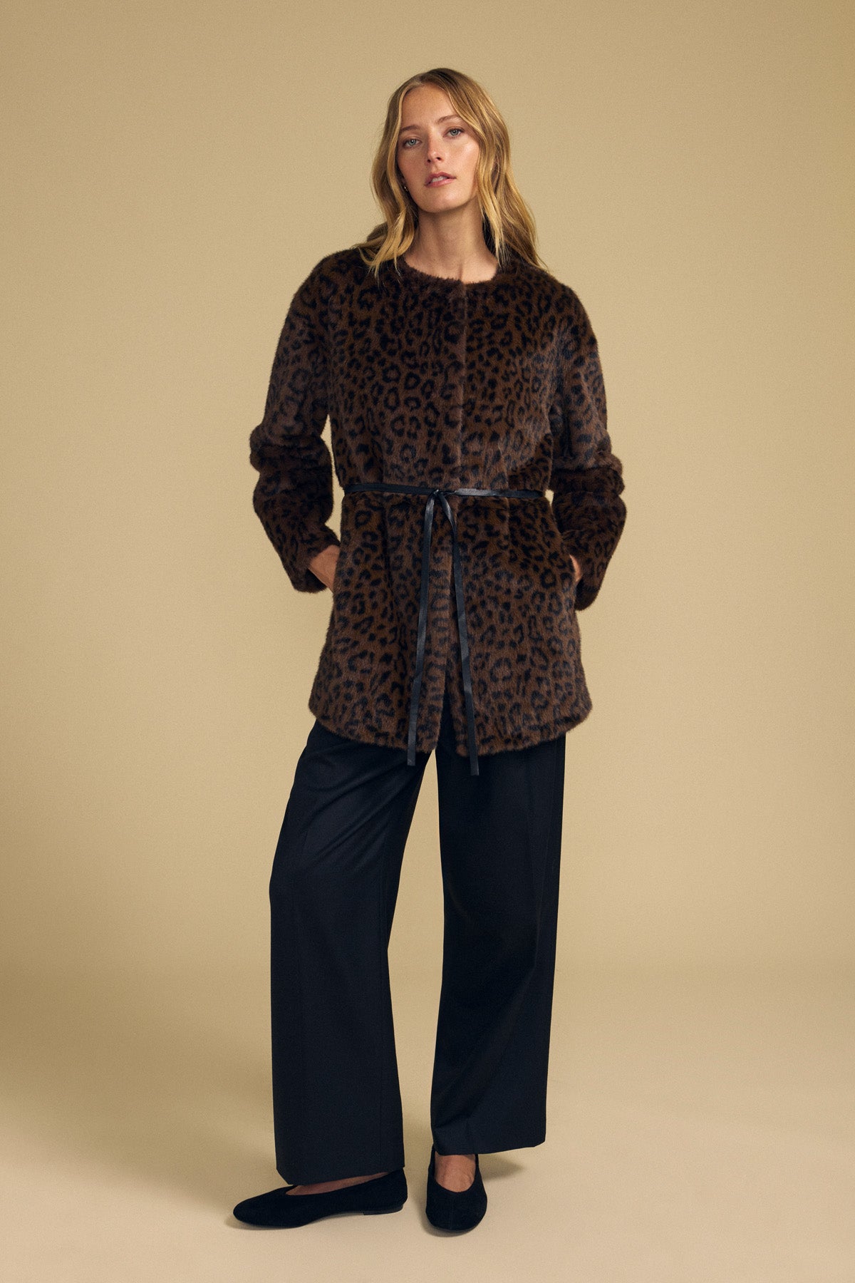 A woman stands against a beige background, wearing the Velvet by Graham & Spencer MARMONT FAUX FUR JACKET—a brown leopard print piece featuring snap button closures and a detachable black belt—paired with black wide-leg pants and black shoes.-42063811510465