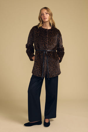 A woman stands against a beige background, wearing the Velvet by Graham & Spencer MARMONT FAUX FUR JACKET—a brown leopard print piece featuring snap button closures and a detachable black belt—paired with black wide-leg pants and black shoes.