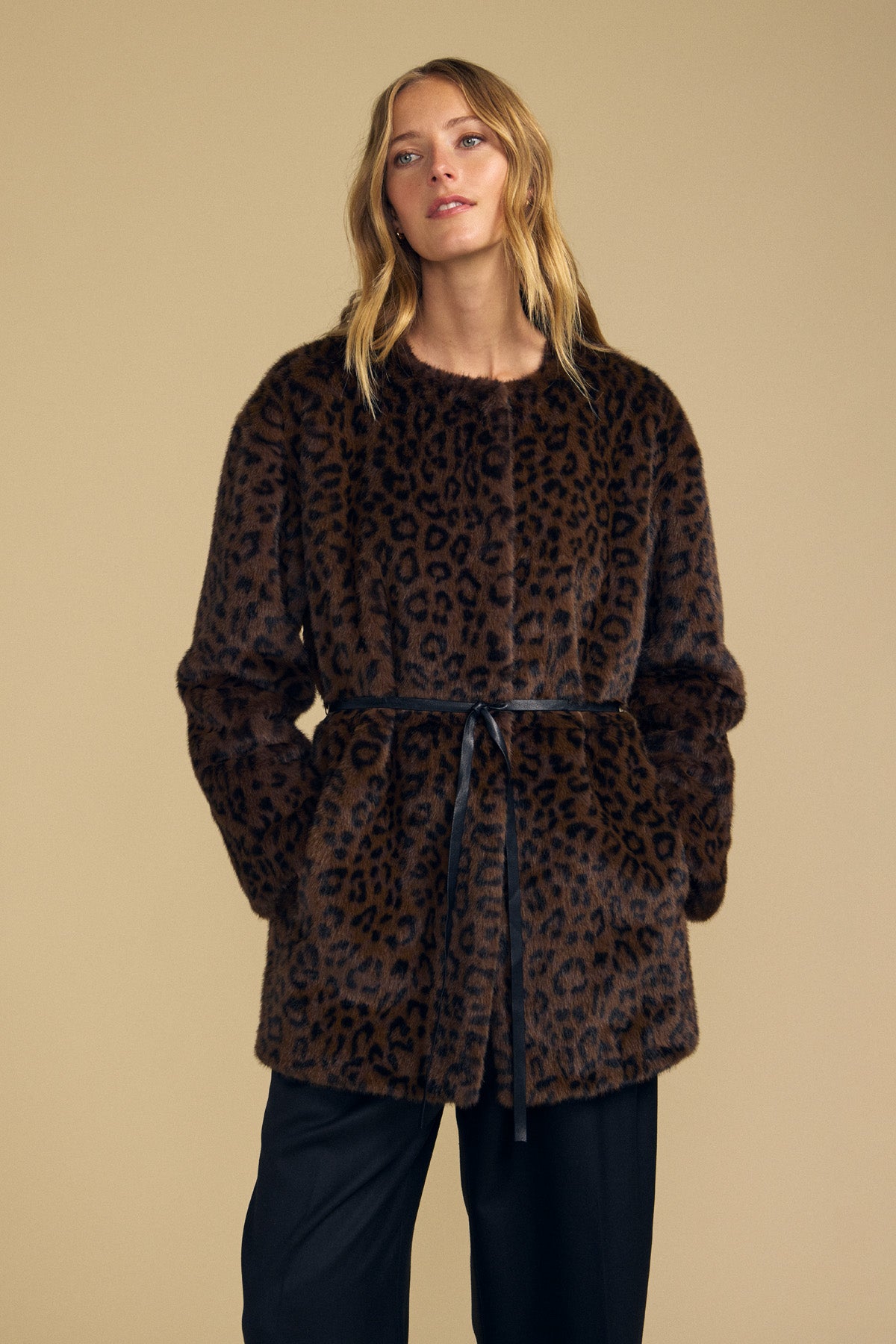 A woman stands against a beige background wearing the Velvet by Graham & Spencer MARMONT FAUX FUR JACKET—a brown leopard print style featuring snap button closures and a detachable black belt—paired with black pants.-42063811576001