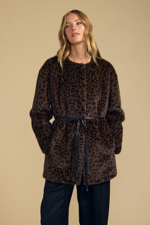 A woman stands against a beige background wearing the Velvet by Graham & Spencer MARMONT FAUX FUR JACKET—a brown leopard print style featuring snap button closures and a detachable black belt—paired with black pants.