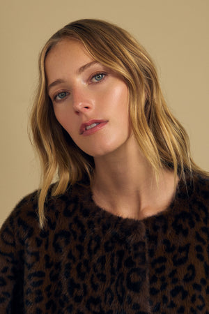 A woman with wavy blonde hair wears the MARMONT FAUX FUR JACKET by Velvet by Graham & Spencer, a leopard print faux fur style with snap button closures, as she gazes slightly to the side against a beige background.