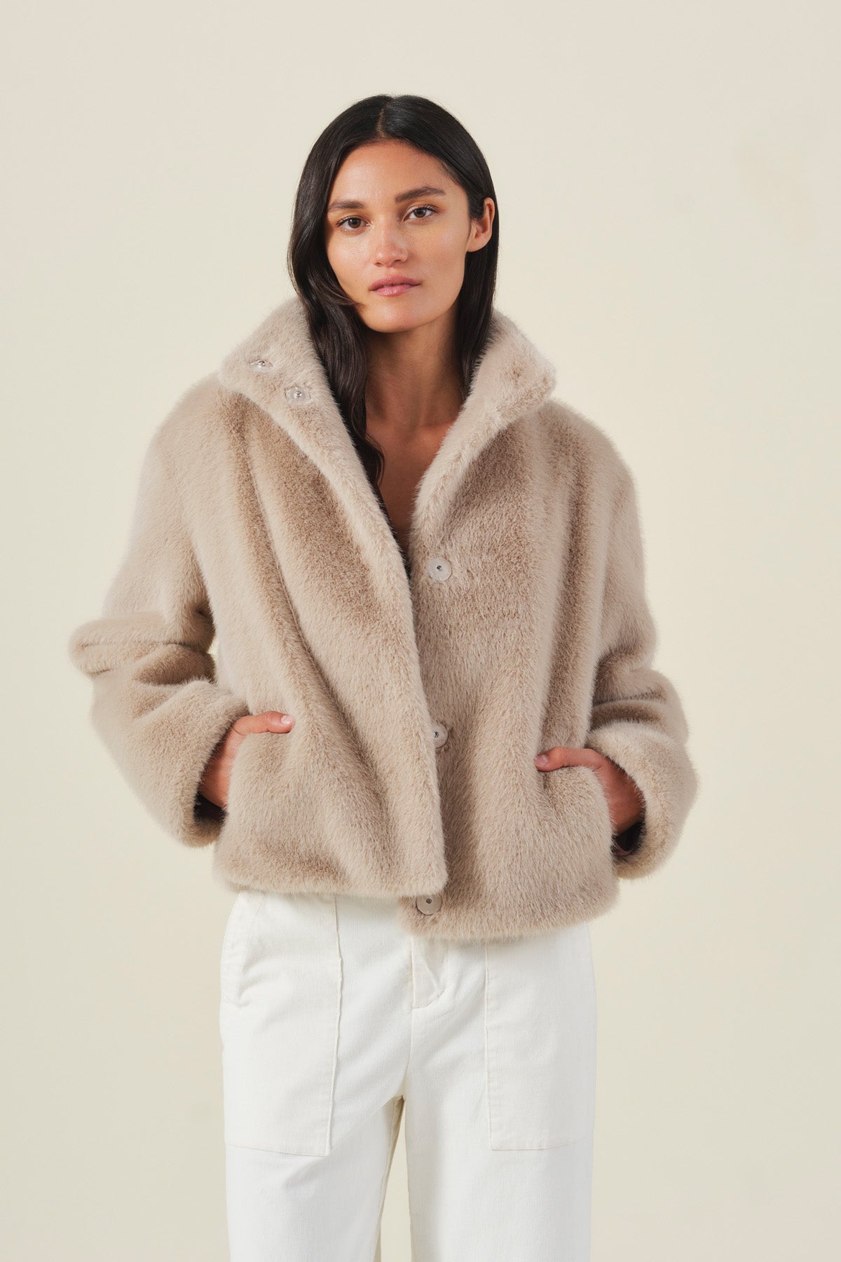 A person stands against a plain background, wearing the Velvet by Graham & Spencer SHEENA FAUX FUR JACKET in beige over white pants, with hands tucked into the jacket pockets and a neutral expression.-42063844769985