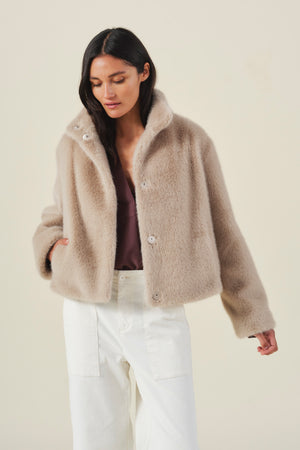 A woman wearing the Velvet by Graham & Spencer SHEENA FAUX FUR JACKET in light brown, featuring a cozy cropped fit, layered over a mauve top and white pants, stands against a plain light background.
