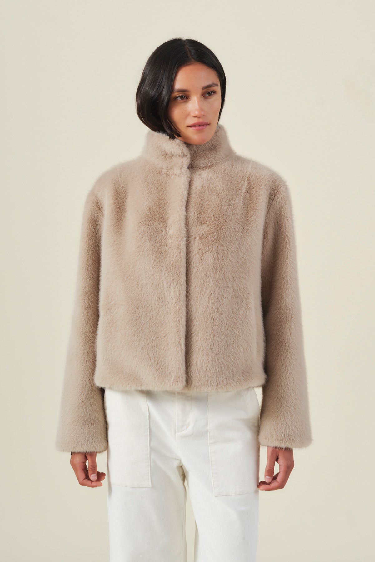 A woman with straight, side-parted dark hair stands against a plain background, wearing the Velvet by Graham & Spencer SHEENA FAUX FUR JACKET in a cozy beige, paired with white pants.