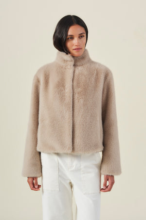 A woman with straight, side-parted dark hair stands against a plain background, wearing the Velvet by Graham & Spencer SHEENA FAUX FUR JACKET in a cozy beige, paired with white pants.