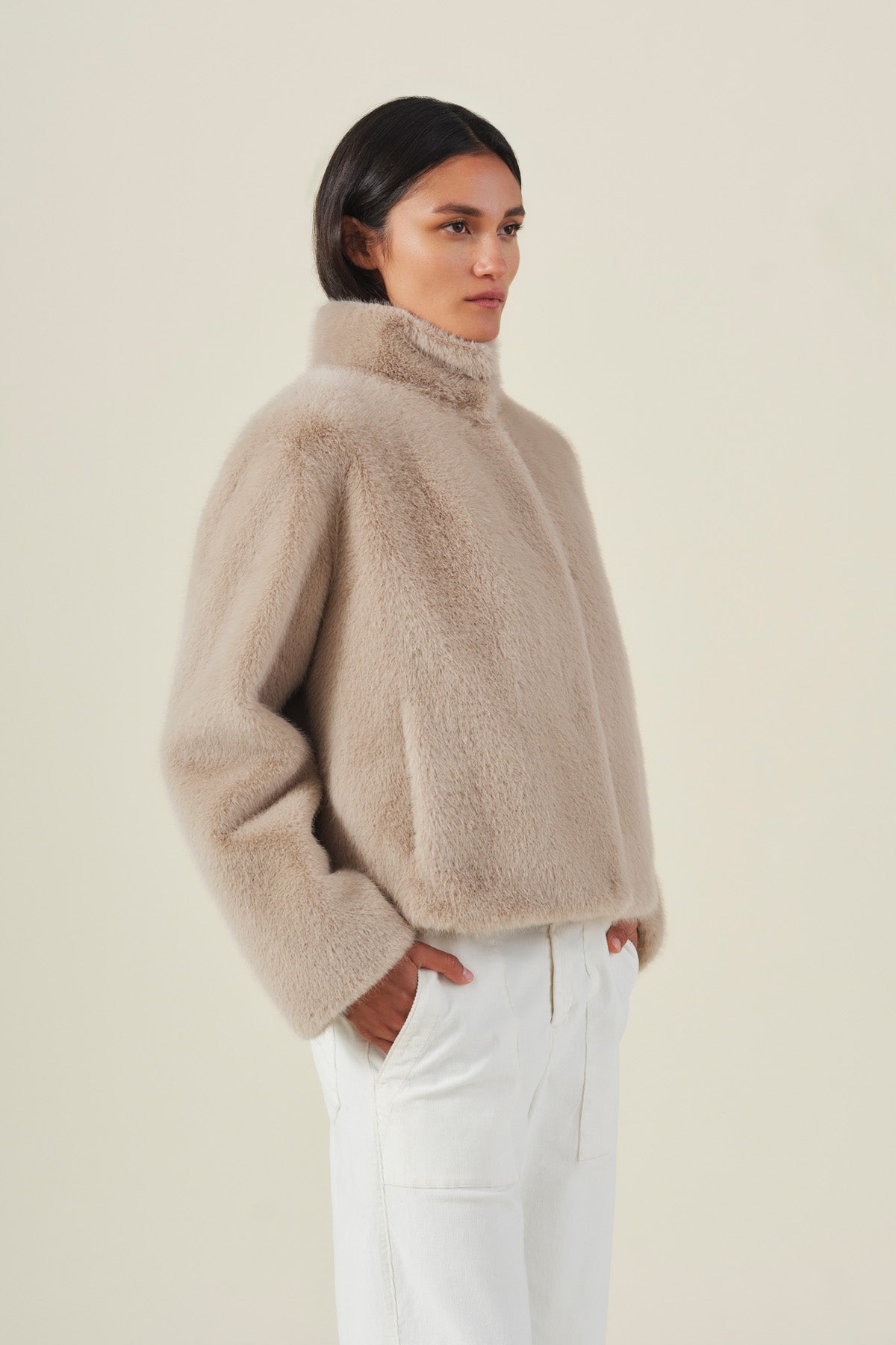 A model wears the SHEENA FAUX FUR JACKET from Velvet by Graham & Spencer—featuring a cozy fit in beige—paired with white pants and stands with hands in pockets against a plain light background.