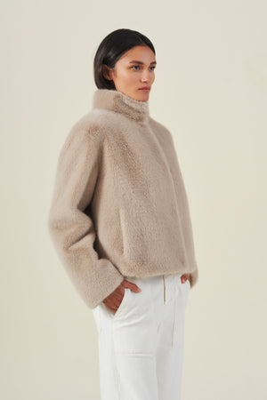 A model wears the SHEENA FAUX FUR JACKET from Velvet by Graham & Spencer—featuring a cozy fit in beige—paired with white pants and stands with hands in pockets against a plain light background.