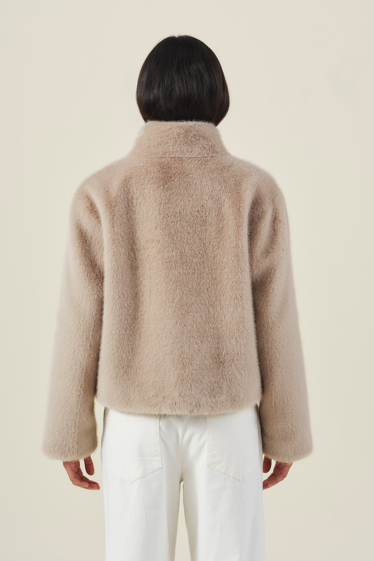 A person is seen from behind, dressed in the Velvet by Graham & Spencer SHEENA FAUX FUR JACKET in cozy beige paired with white pants, set against a plain light background.