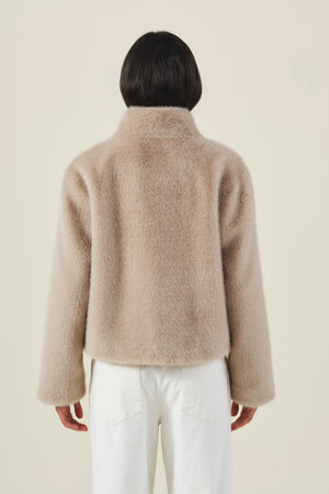 A person is seen from behind, dressed in the Velvet by Graham & Spencer SHEENA FAUX FUR JACKET in cozy beige paired with white pants, set against a plain light background.