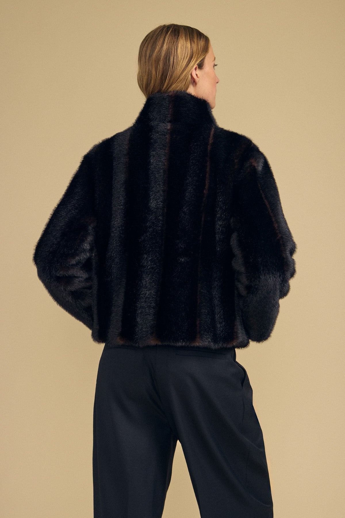 A person with light hair is shown from the back wearing the SHEENA FAUX FUR JACKET by Velvet by Graham & Spencer, styled in a cozy fit with black pants, standing against a plain beige background.