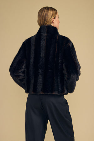 A person with light hair is shown from the back wearing the SHEENA FAUX FUR JACKET by Velvet by Graham & Spencer, styled in a cozy fit with black pants, standing against a plain beige background.