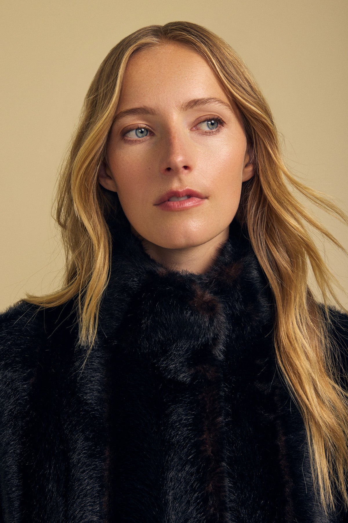 A woman with long blonde hair wears the SHEENA FAUX FUR JACKET by Velvet by Graham & Spencer, featuring a cozy fit in black, as she looks slightly to the side against a plain beige background.