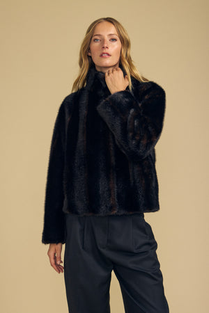 Against a beige background, a person wears the SHEENA FAUX FUR JACKET by Velvet by Graham & Spencer—a dark cropped faux fur style—paired with black pants, with one hand gently touching the collar for a cozy fit.