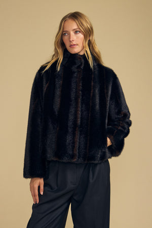 A woman wearing the SHEENA FAUX FUR JACKET by Velvet by Graham & Spencer, featuring a cozy fit in a dark hue, pairs it with black trousers and stands against a plain beige background, gazing slightly to the side.