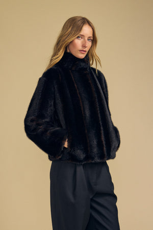 A woman wearing the Velvet by Graham & Spencer SHEENA FAUX FUR JACKET in black, paired with black pants, stands against a plain beige background and looks at the camera.