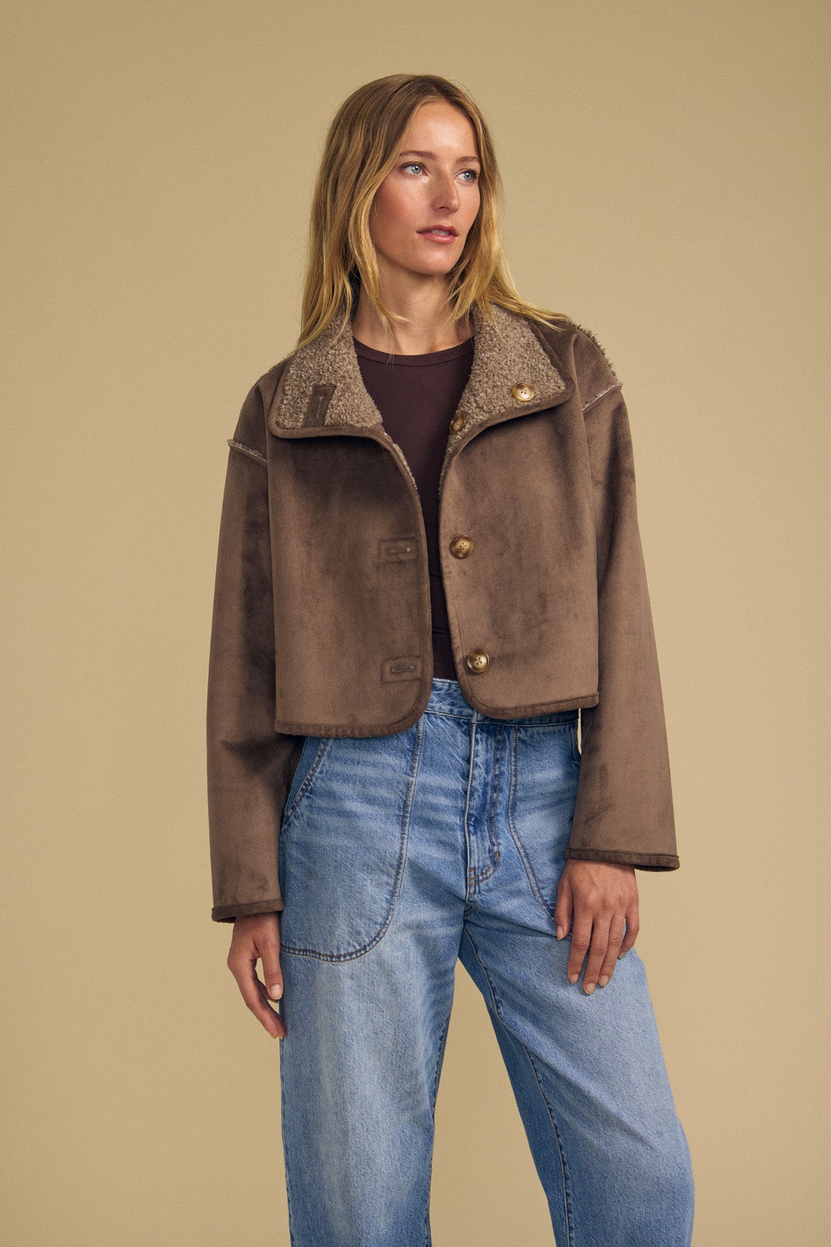 A woman stands against a beige background wearing the ESME FAUX SHERPA REVERSIBLE JACKET by Velvet by Graham & Spencer—a brown faux suede cropped jacket with a shearling collar—styled over a dark top and high-waisted blue jeans.-41971005161665