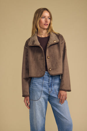 A woman stands against a beige background wearing the ESME FAUX SHERPA REVERSIBLE JACKET by Velvet by Graham & Spencer—a brown faux suede cropped jacket with a shearling collar—styled over a dark top and high-waisted blue jeans.