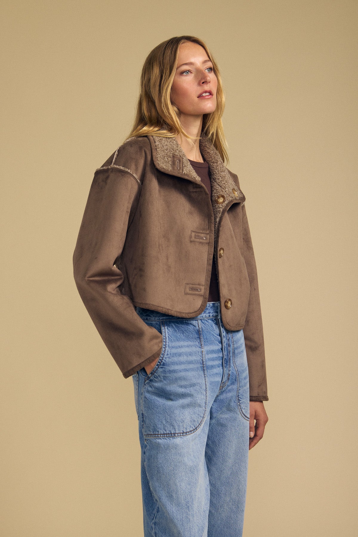 A woman stands against a beige background wearing the Velvet by Graham & Spencer ESME FAUX SHERPA REVERSIBLE JACKET, featuring large buttons, plush faux sherpa lining, and soft faux suede on the exterior, paired with blue high-waisted jeans.-41971005194433