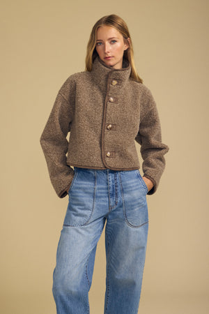 A person wearing the ESME FAUX SHERPA REVERSIBLE JACKET by Velvet by Graham & Spencer and blue wide-leg jeans stands with hands in pockets against a beige background.
