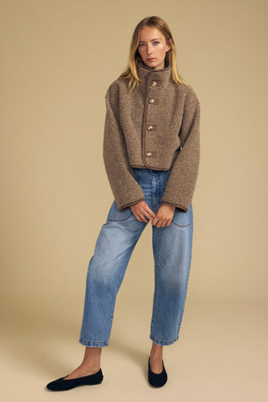 A woman stands against a beige background, wearing the ESME FAUX SHERPA REVERSIBLE JACKET by Velvet by Graham & Spencer, paired with loose blue jeans and black flats.